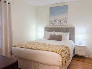 Ein ordentlich bezogenes Bett mit einer gemusterten Tagesdecke, ein Nachttisch mit einer Lampe und ein Strandbild an der Wand.