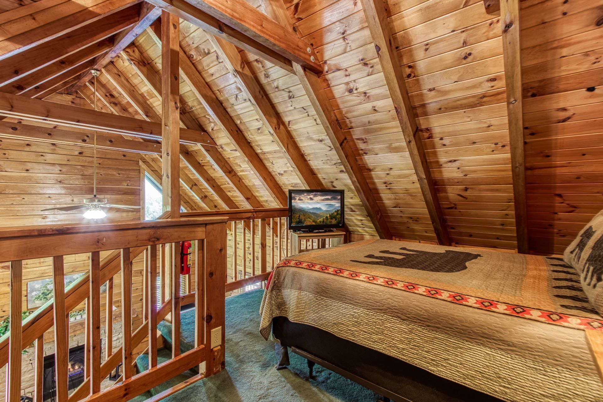 Een slaapkamer in een houten blokhut met een schuine plafond, een bed met een bedspread met dessin, een wandtelevisie en een houten leuning.