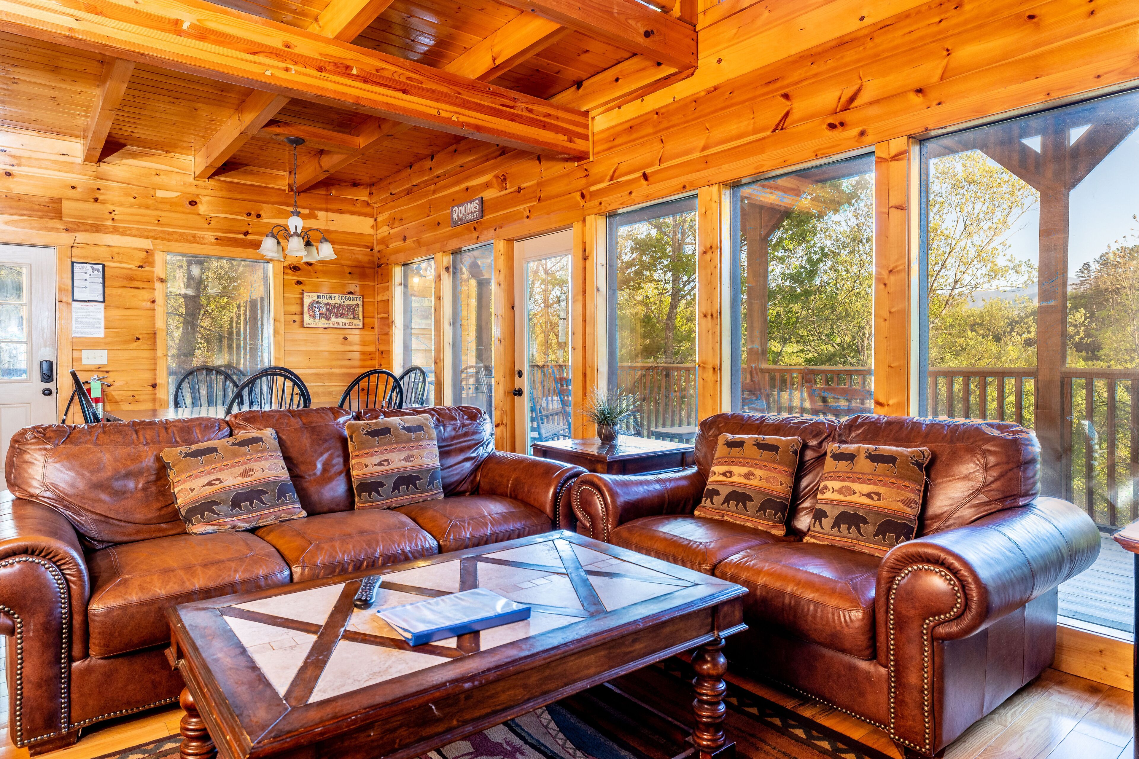 Interior de una cabaña de madera con sofá de cuero, una mesa de centro de madera y ventanas amplias que dan al exterior.