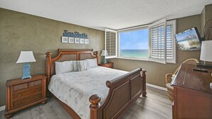 Ein Schlafzimmer mit einem hölzernen Bett, einem Nachttisch mit Lampe, einem Schreibtisch mit Computer und einem Fenster mit Meerblick.