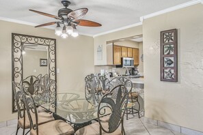 A dining area with a glass table and metal chairs, a ceiling fan, and a kitchen in the background. - 🌊 Islander #703 – Stylish Beachfront Escape with Stunning Gulf Views 🏖️✨ (Destin)