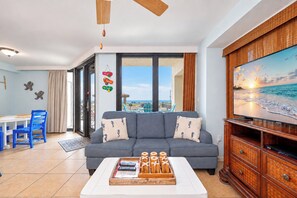 Una sala de estar con un sofá, una mesa de centro y un televisor. Se ve la playa a través de las ventanas.