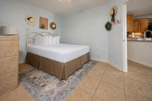 A bedroom with a bed, a dresser, a rug, and a kitchen visible through an open door. - GROUND FLOOR PREMIUM! Phoenix III ~ Corner Condo on the BEACH! (Orange Beach)