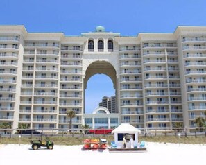 A large hotel with multiple balconies, a central arched entrance, and a beach area with a small kiosk and lounge chairs.