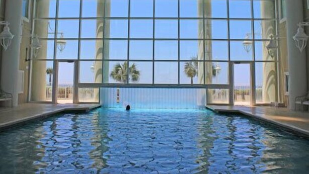 A large indoor swimming pool with clear blue water, surrounded by large windows offering a view of palm trees and a clear sky.