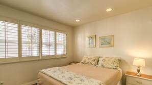 A bedroom with a bed, a nightstand, a lamp, two framed pictures on the wall, and a window with blinds. - 219D: Spacious Two-Level Home, Mountain Views, Fireplace, Close to Beach, BBQ - (Avalon)