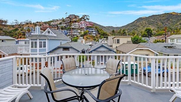 A balcony with a round table and chairs, overlooking a hillside with multi-story buildings. - 219D: Spacious Two-Level Home, Mountain Views, Fireplace, Close to Beach, BBQ - (Avalon)