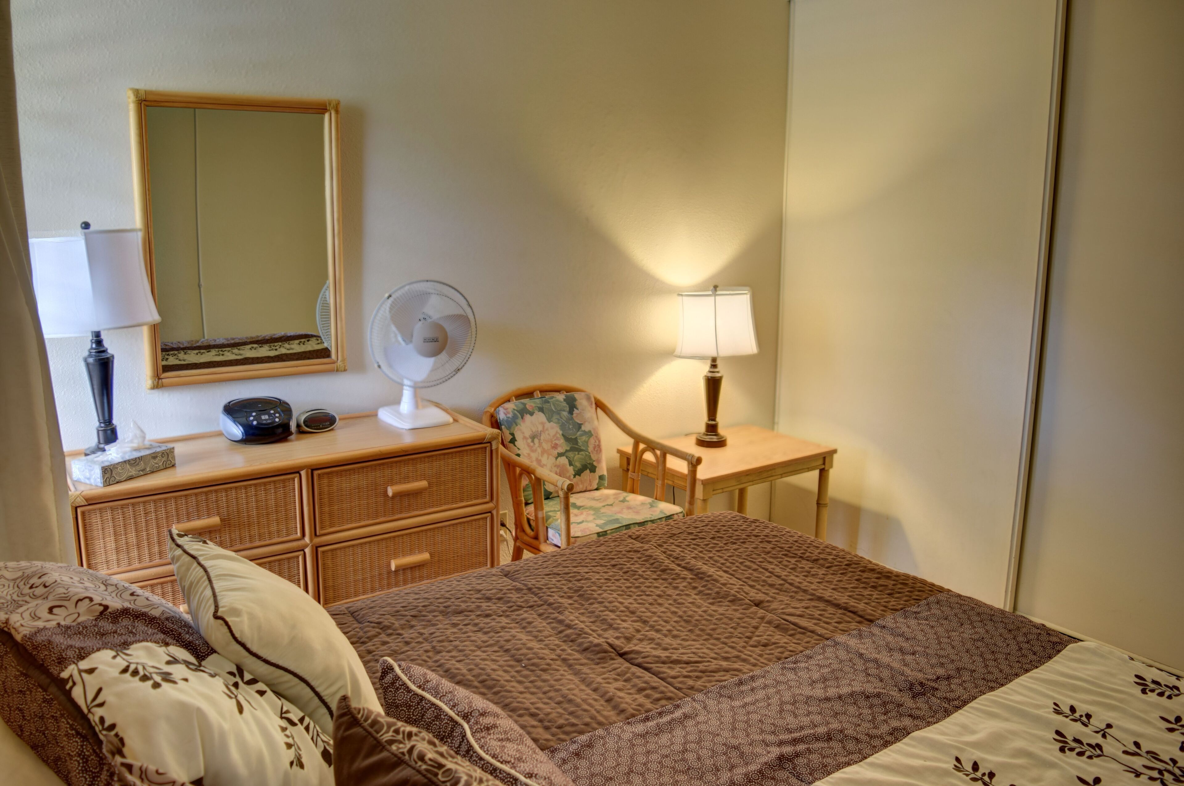 A bedroom with a bed, a dresser, a chair, a table, a lamp, a fan, and a mirror.