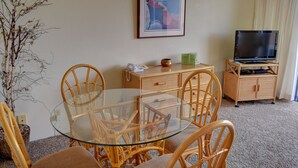 Un soggiorno con un tavolo da pranzo in vetro circondato da sedie in vimini, una televisione su un mobile e un quadro appeso alla parete.