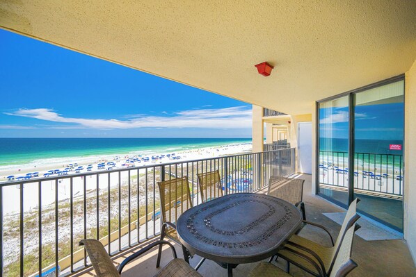 A balcony with a view of the beach, a table, chairs, and a railing.