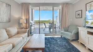 A modern living room with a sofa, a coffee table, a TV, and a balcony with a view of the sky and greenery. - Waterfront 1BR + Bunk Alcove at The Wharf • Sleeps • Amazing Views • Oasis Pool (Orange Beach)