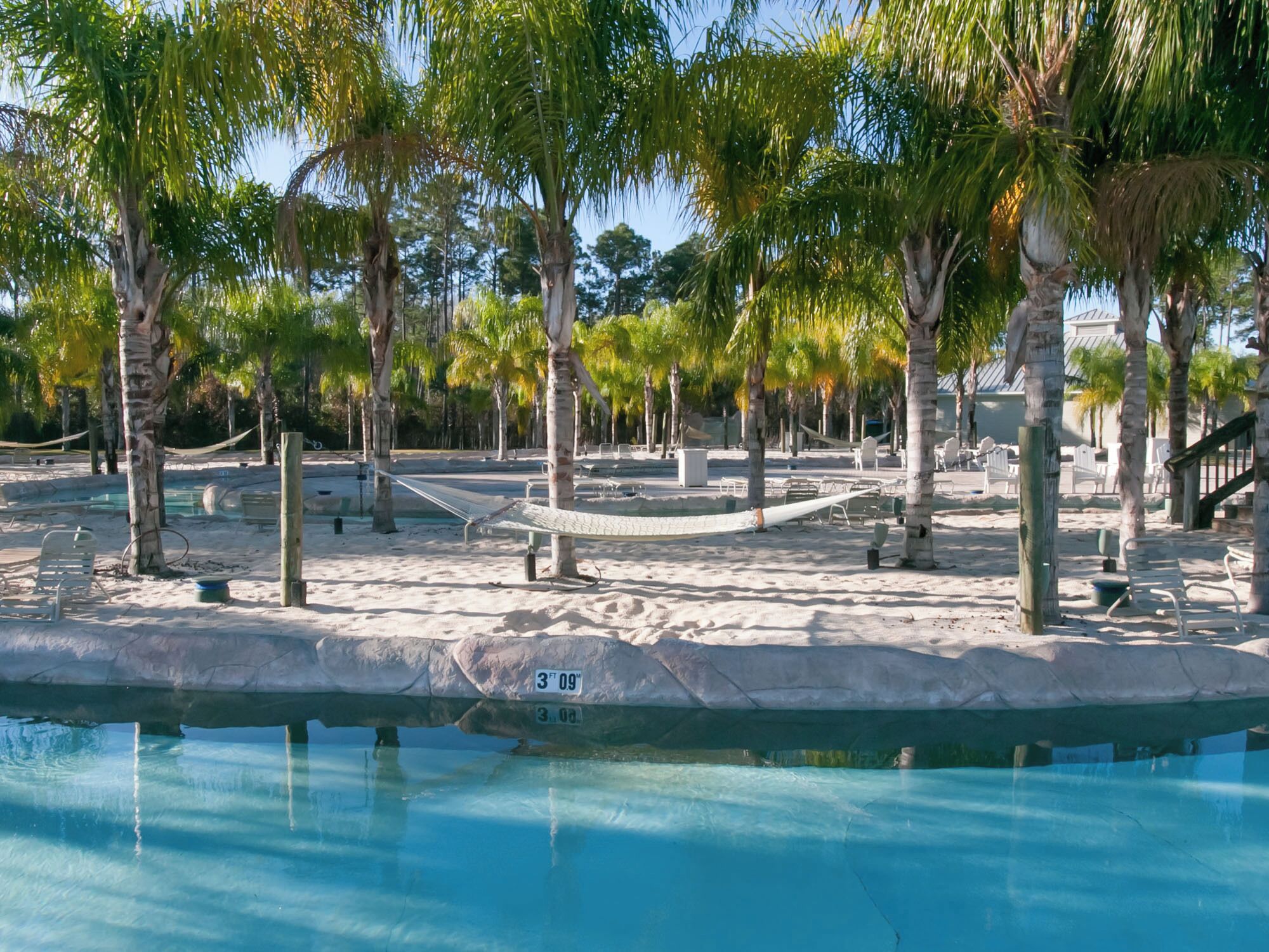 Un espace piscine avec une plage de sable, des palmiers et un hamac.