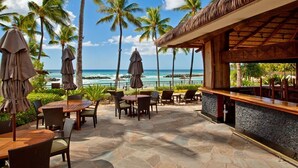 Un patio frente a la playa con mesas y sillas, una zona de bar y palmeras.