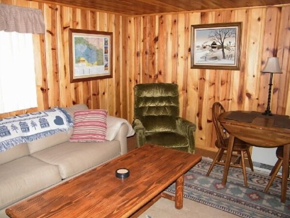 A cozy room with wooden walls, a sofa, a green armchair, a wooden table, and a framed map on the wall. - With2Beez Rustic Forest Cabin (Greer)
