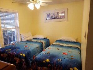 Ein Zimmer mit zwei Betten, einem Deckenventilator, einem Fenster mit Jalousien und einem gerahmten Bild an der Wand.