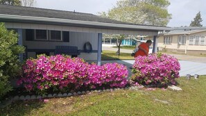 Ein Haus mit überdachter Veranda, einer Gartenschlauchaufrolle und einem gepflegten Garten mit rosa Blüten.