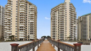 A wooden boardwalk leading to high-rise buildings with balconies, a clear sky, and sandy beach.