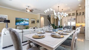 A dining area with a table set for six, surrounded by chairs. There is a sofa and a television in the background.