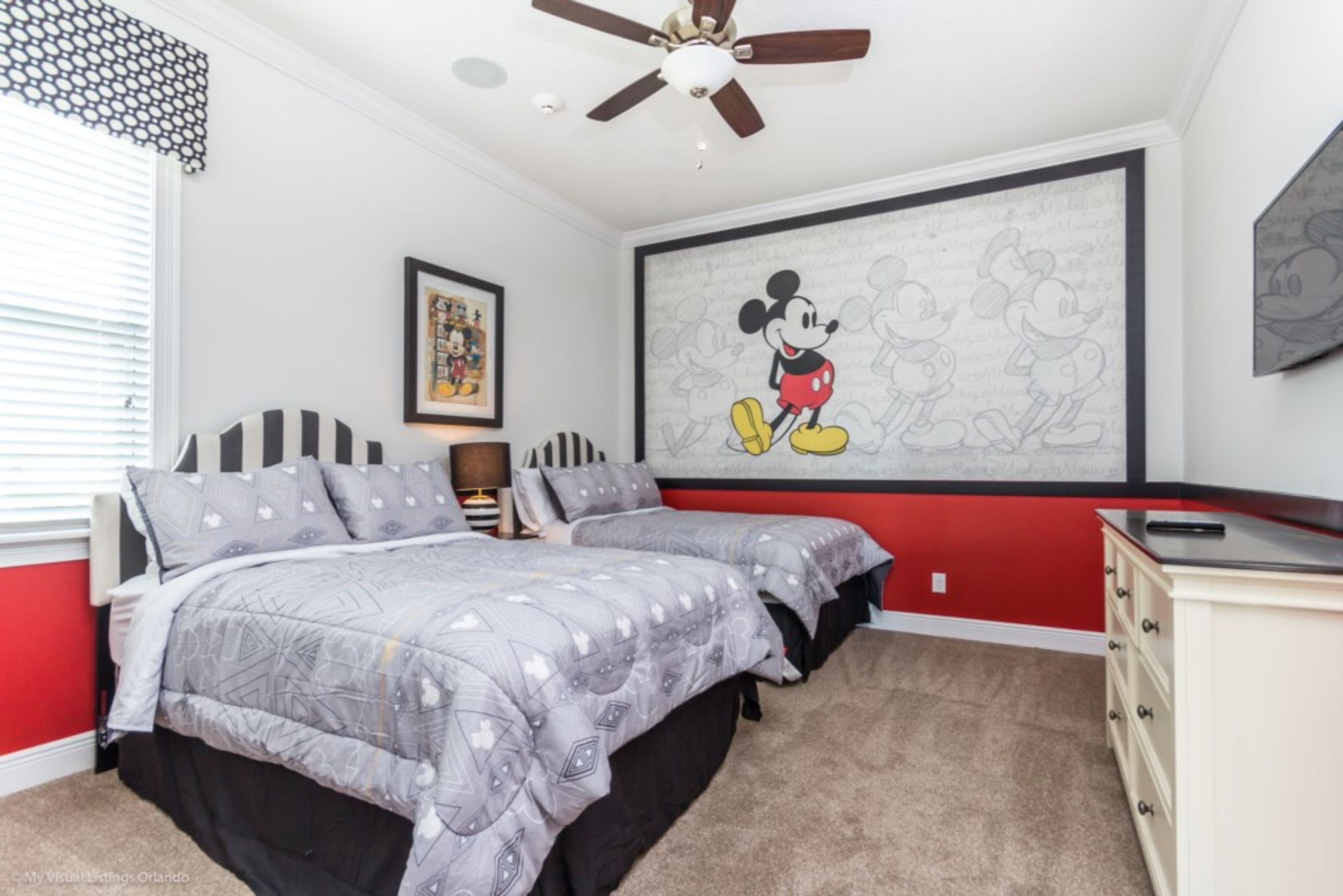 A bedroom with two beds, a ceiling fan, a large Mickey Mouse mural, and a dresser.