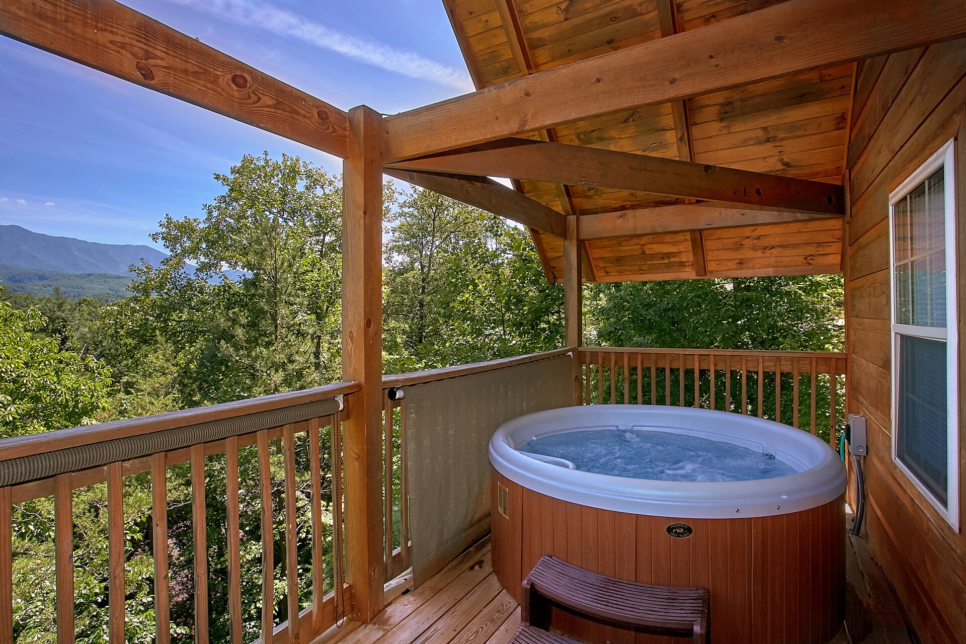 Una terraza de madera con un jacuzzi, barandillas de madera y vistas a montañas y árboles.
