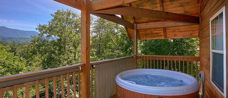 Una terraza de madera con un jacuzzi, barandillas de madera y vistas a montañas y árboles.