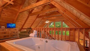 Interior de una cabaña de madera con una bañera grande, vigas de madera, un televisor y vistas al verdor exterior.