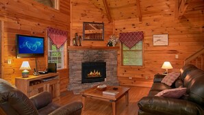 Un interior de cabaña acogedor con chimenea de piedra, mobiliario de cuero, televisor y paredes de madera.
