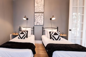 Una habitación con dos camas, cada una con una almohada de estampado blanco y negro, una mesita de noche con lámpara y una obra de arte colgada en la pared.