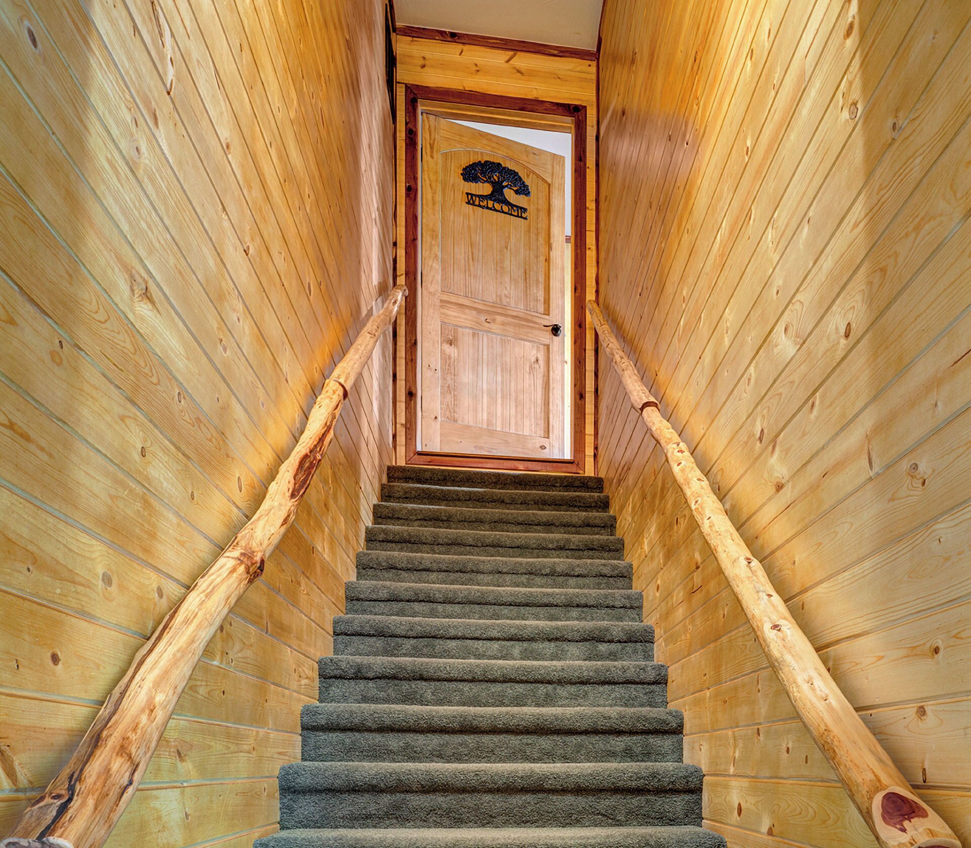 A wooden staircase with a carpeted landing leading to a wooden door with a welcome sign.