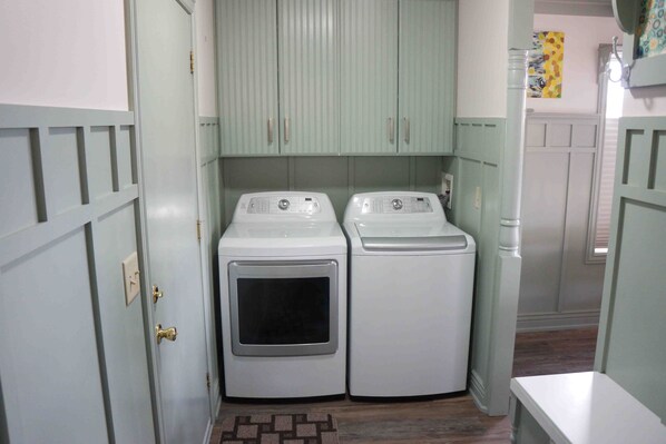 Une buanderie équipée de deux machines à laver et d’un sèche-linge, avec des placards vert clair et un tapis à motifs sur le sol.