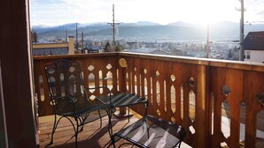 Un balcon doté d’une balustrade en bois et de chaises en métal, offrant une vue sur un paysage urbain avec des montagnes à l’horizon.