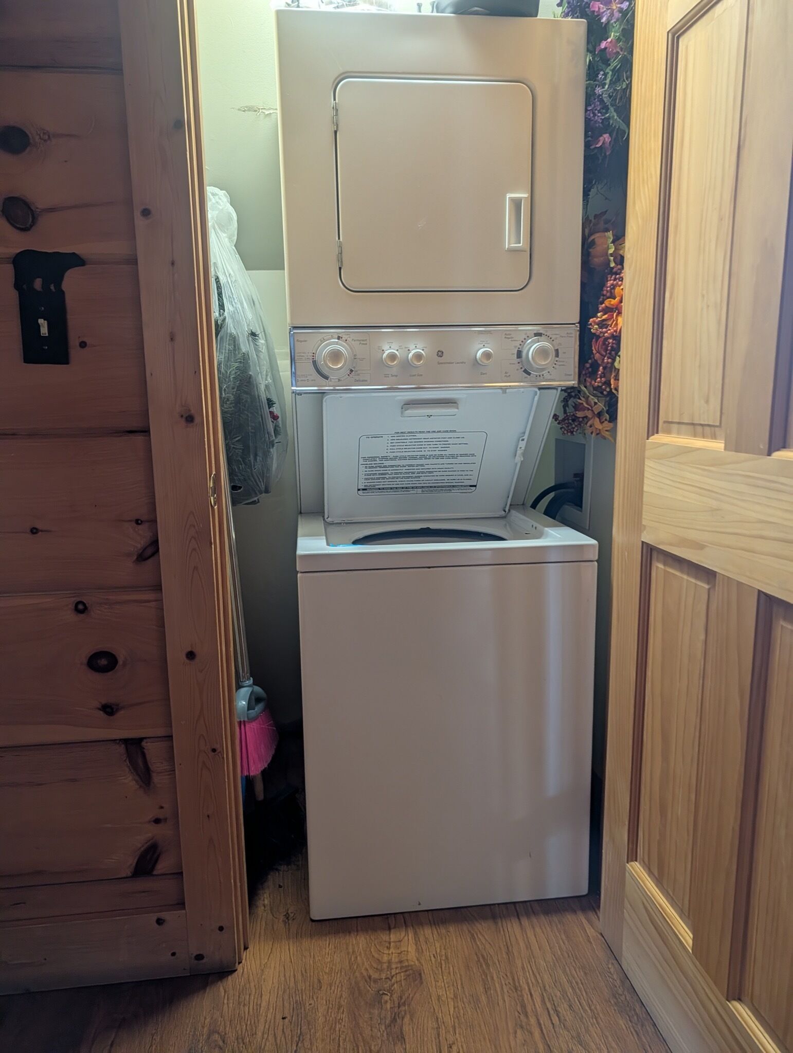 Une petite buanderie équipée d’un lave-linge et d’un sèche-linge superposés, de meubles en bois et d’une petite poubelle rose.
