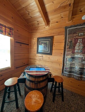 A wooden cabin interior with a wooden ceiling, a window with plaid curtains, a round table with a flag design, two stools, and a tapestry on the wall.