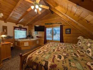 A cozy attic bedroom with a wooden ceiling, a large bed with a patterned quilt, a built-in bathtub, and a television mounted on the wall.