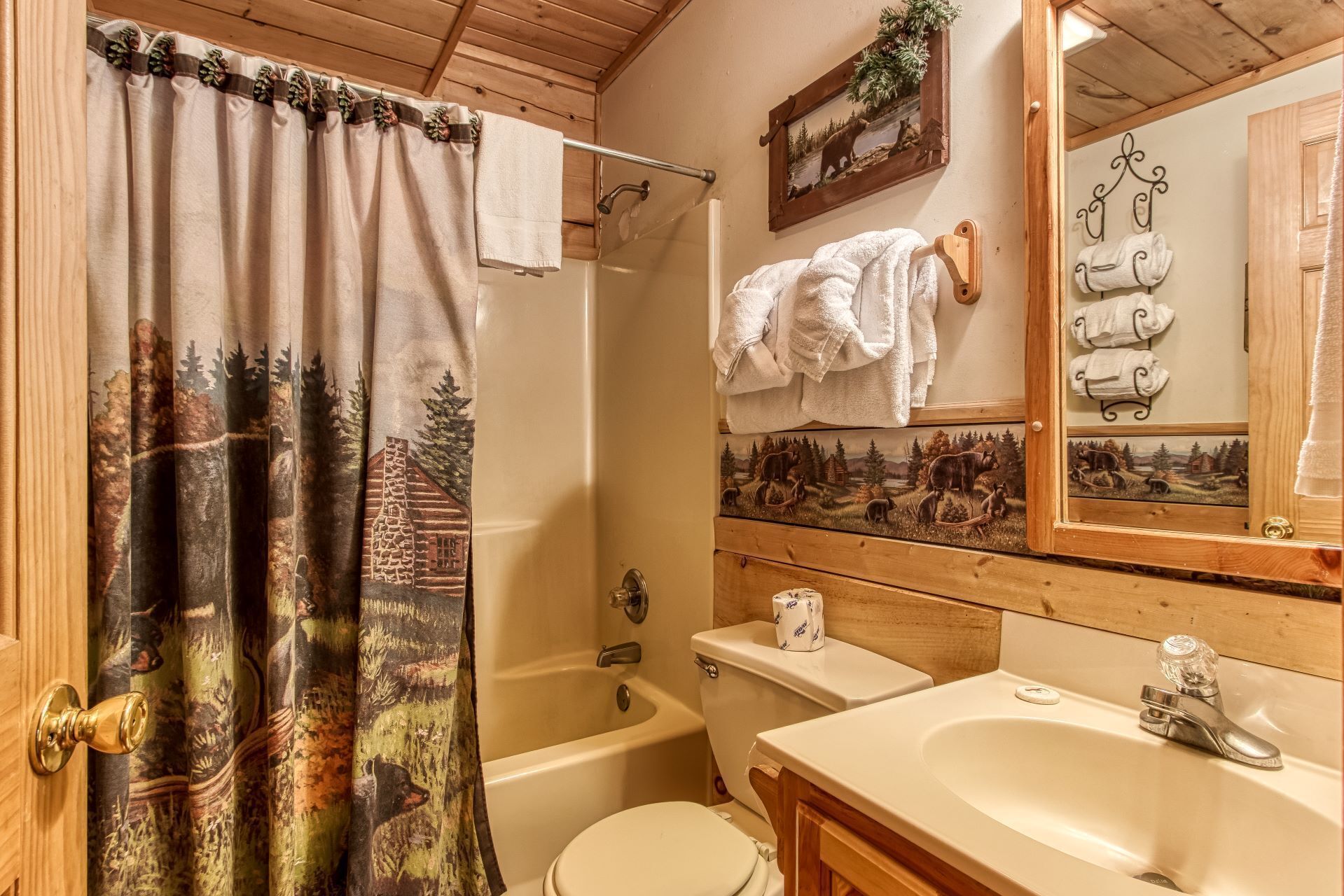 A bathroom with a shower curtain depicting a forest scene, a toilet, a bathtub, a sink, and a mirror with multiple towel hooks.