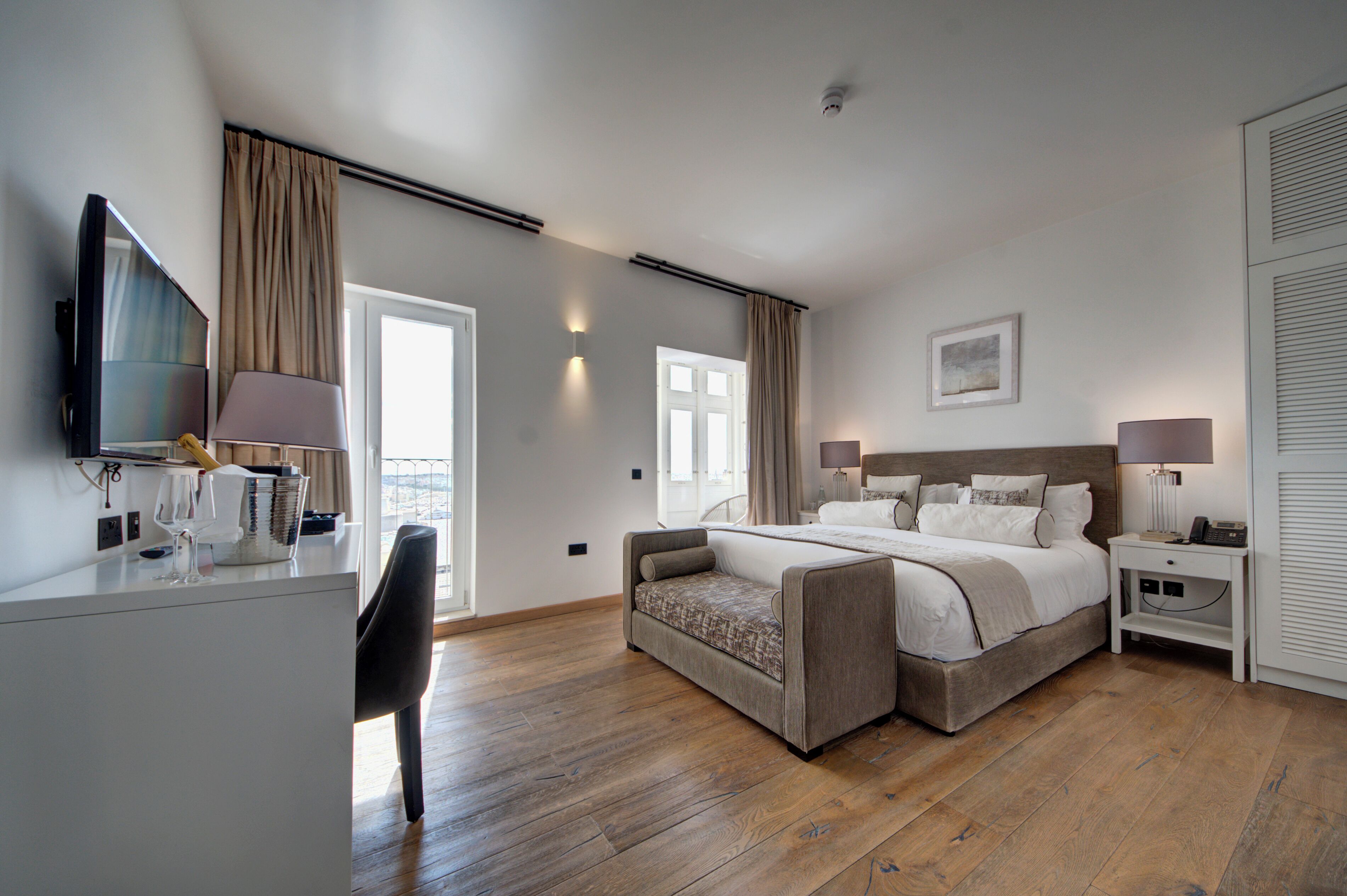 A hotel room with a bed, a desk with a TV, a chair, a bedside table with lamps, and a window.