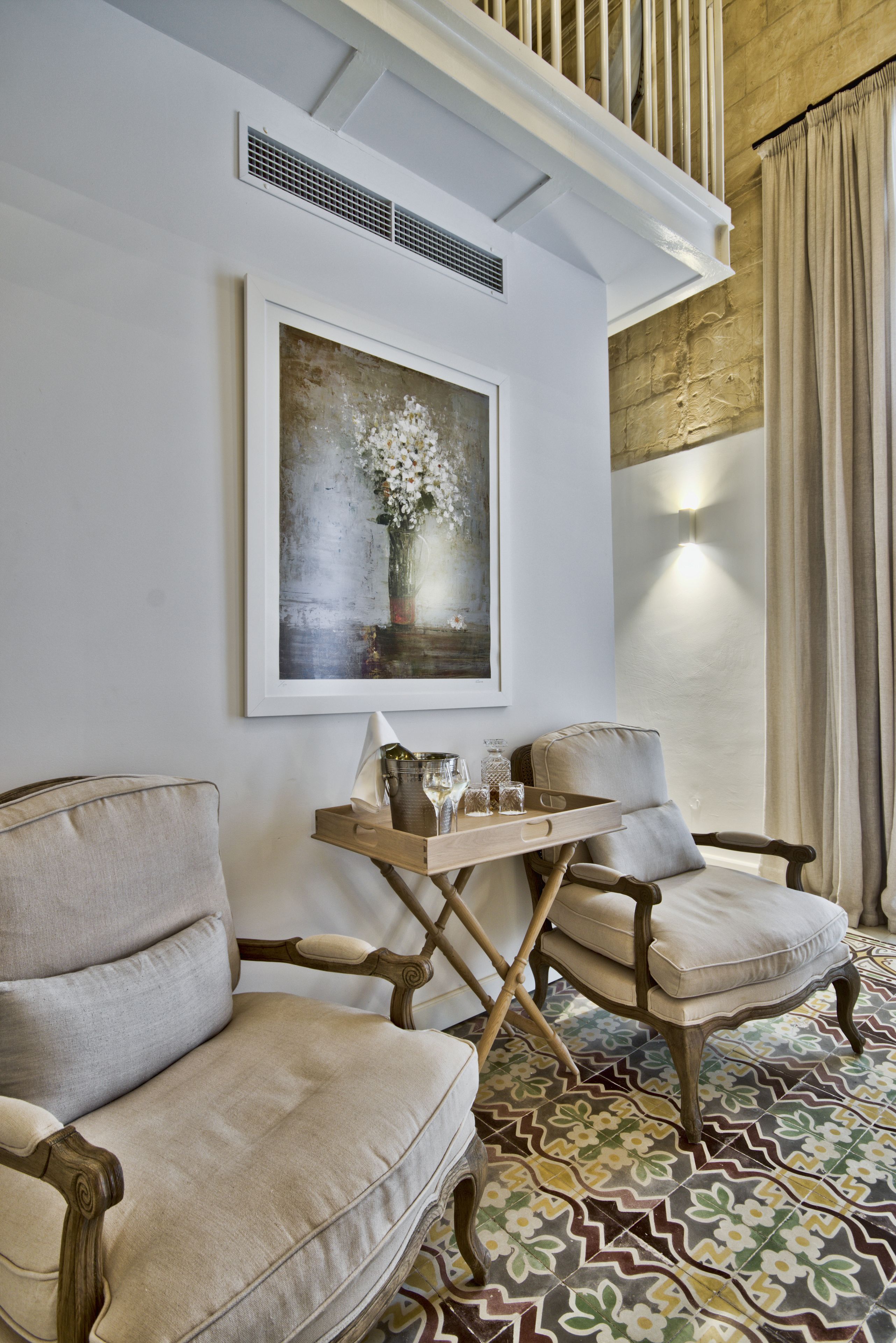A room with two upholstered chairs, a wooden side table with glassware, and a framed painting on the wall.