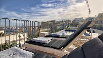 A rooftop lounge area with lounge chairs and a pool, overlooking a cityscape.