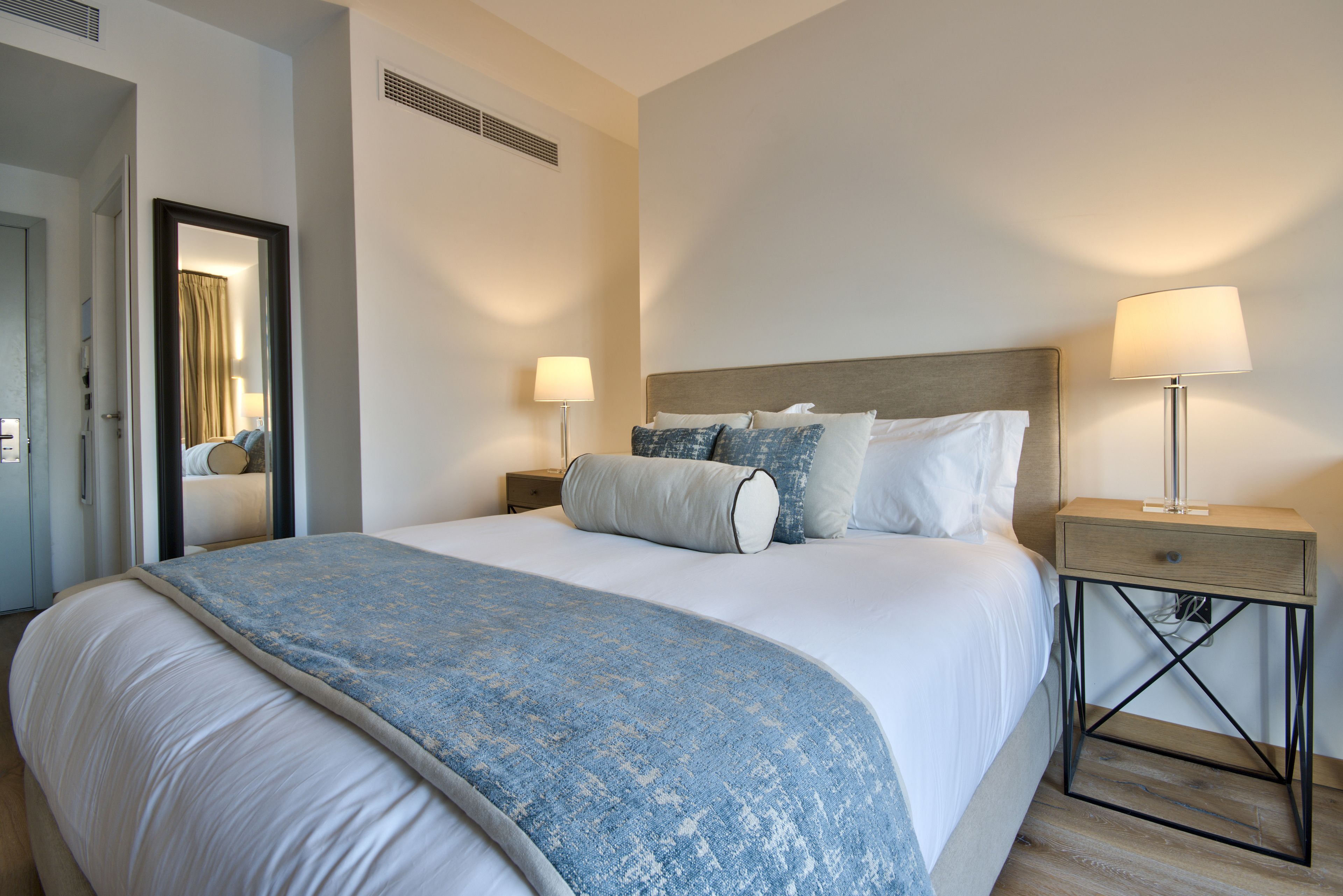 Ein ordentlich bezogenes Bett mit blauer Decke, ein Nachttisch mit Lampe und ein Spiegel an der Wand.