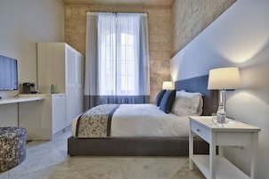 Ein modernes Hotelzimmer mit Bett, Nachttisch, Lampe und Blick auf die Stadt durch ein großes Fenster.