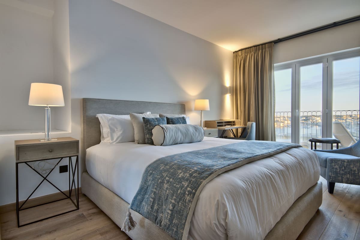 A modern hotel room with a large bed, two bedside tables, a desk, and a view of the city through large windows.
