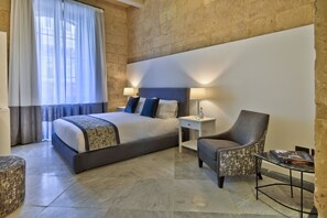 Quarto de hotel moderno com uma cama grande, duas luminárias de cabeceira, uma cadeira, uma mesinha e uma janela com cortinas.