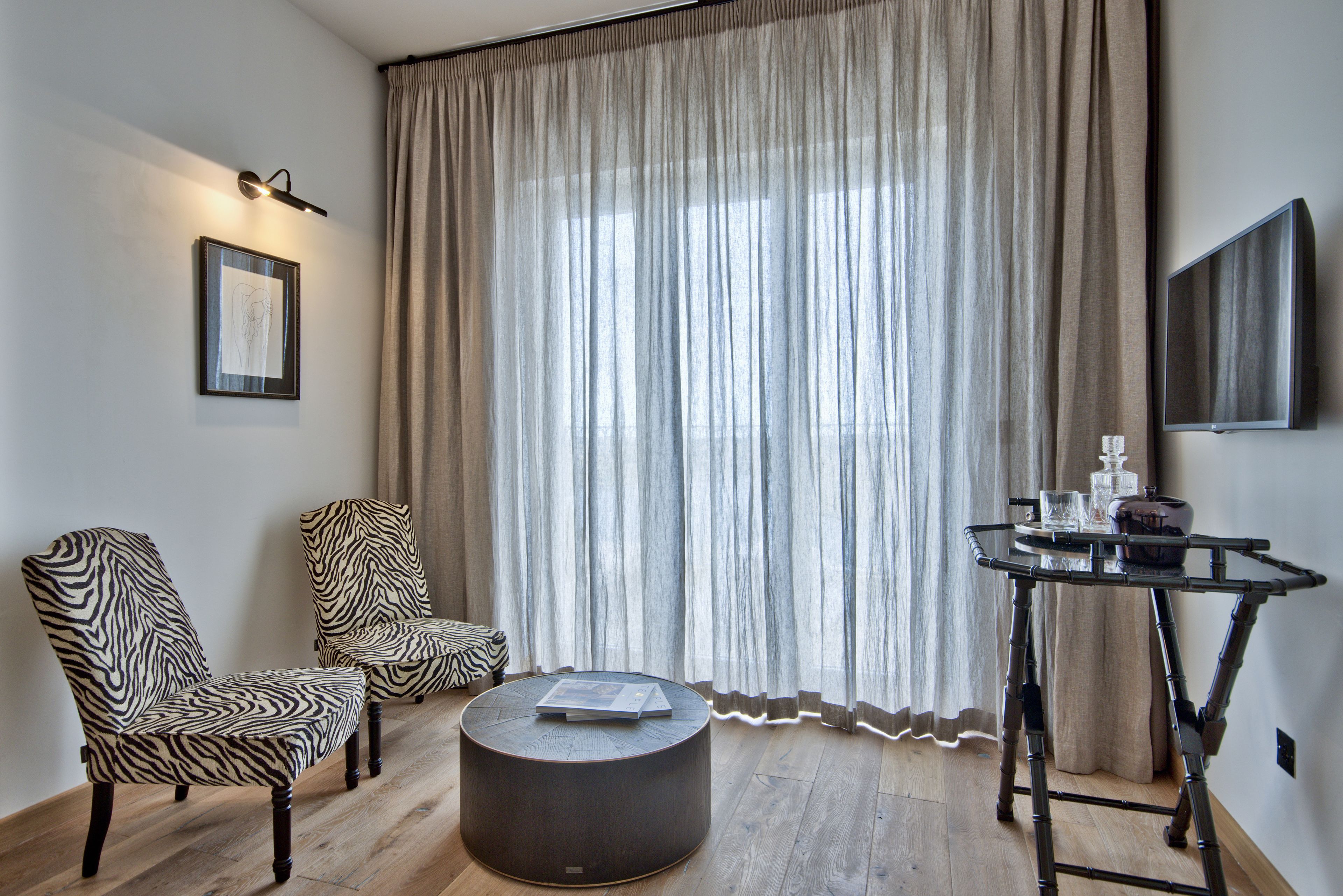 A room with two zebra-patterned chairs, a round coffee table, a small side table with a glass and a vase, and a large window with sheer curtains.