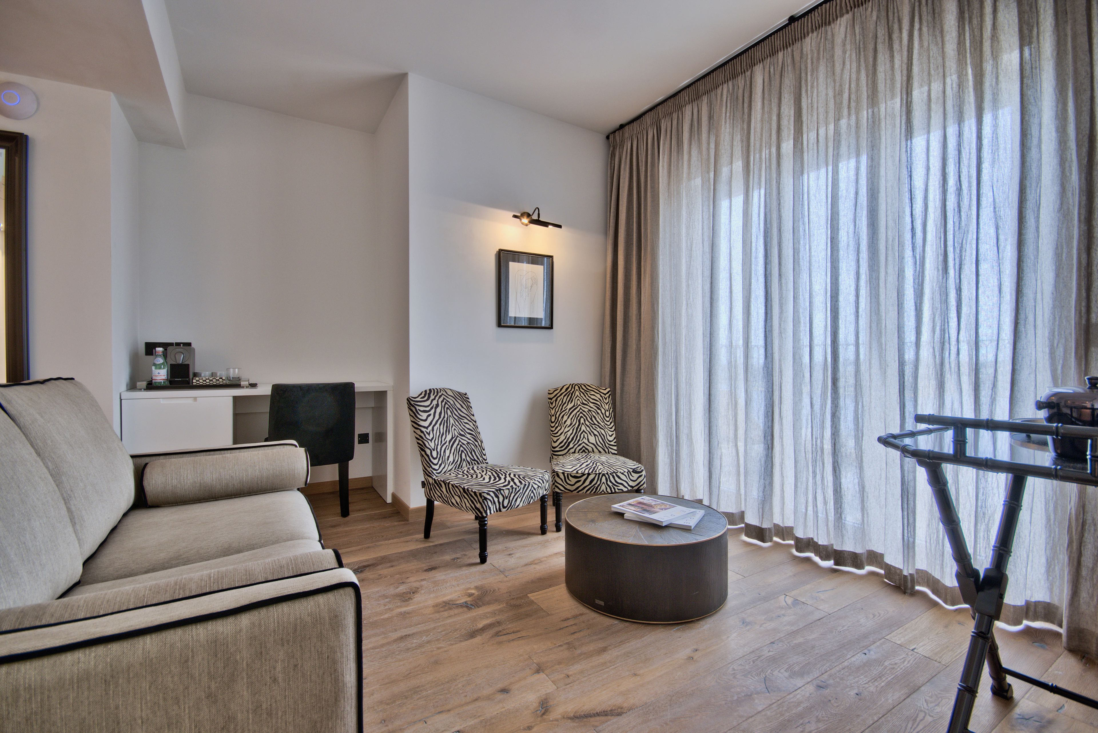 Una sala de estar moderna con un sofá, dos sillones con estampado de cebra, una mesa de centro redonda y una mesa de comedor con una silla. Se observa un cuadro en la pared y una pequeña zona de kitchenette al fondo.
