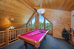 A wooden room with a pool table, a ceiling fan, and a view of trees through a window.