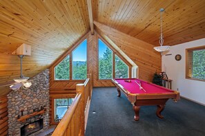 A wooden interior with a pool table, stone fireplace, and large windows offering a view of the outdoors.