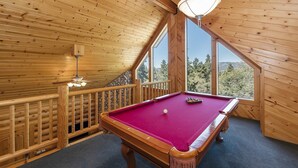 A wooden room with a pool table, a ceiling fan, and a large window offering a view of trees and mountains.
