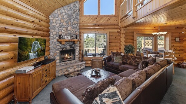 A log cabin interior with a stone fireplace, a large sofa, a television, and a balcony with a view of trees.