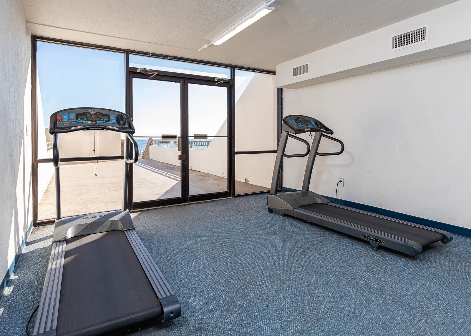 Een fitnessruimte met twee loopbanden, een grote schuifglazen deur die leidt naar een buitenruimte, en een witte muur met een ventilatie rooster.