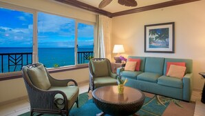 A balcony with a view of the ocean, a green sofa, two wicker chairs, a round table, and a ceiling fan.